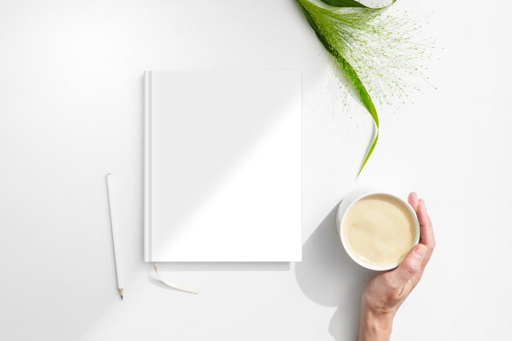 The notebook is on a white table with a plant, pens and a cup of coffee. A hand holds a cup.