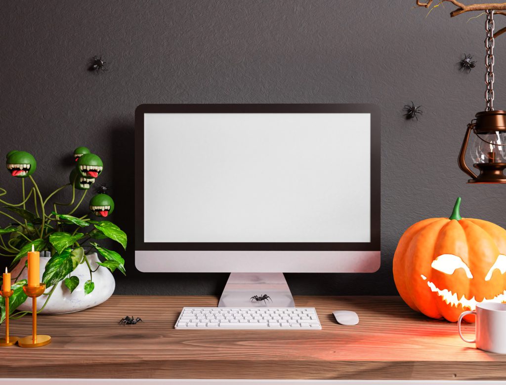 iMac computer on the table. Next to the computer is a pumpkin. There are candles, a scary plant, spiders and a cup on the table.