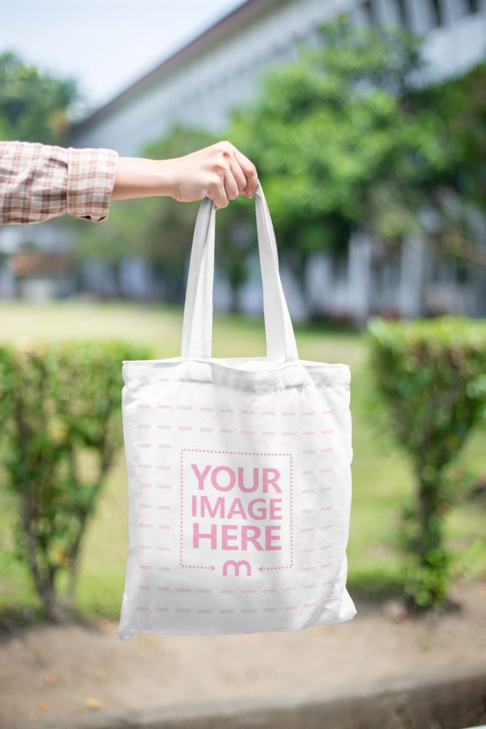 A person is holding a tote bag. The human body is not visible, only the hands. In the background is a garden and bushes and a large house.