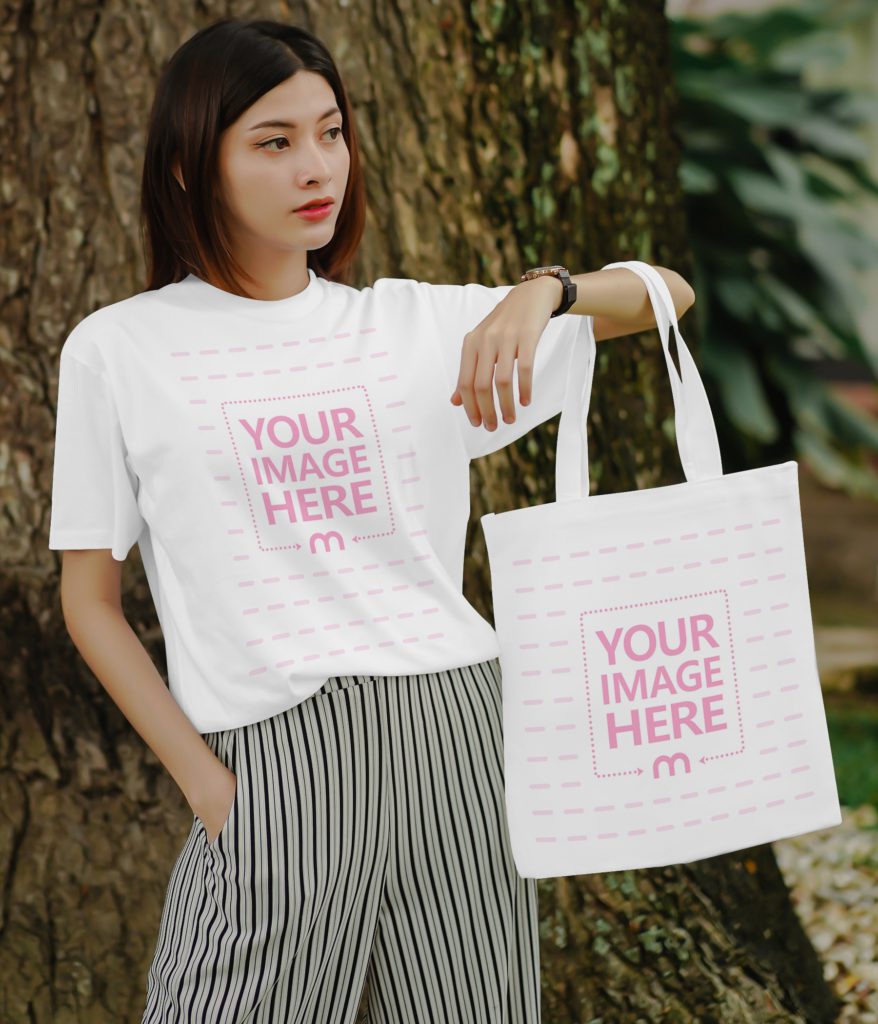 A confident woman stands with her hands in her pockets and holding a tote bag in the other hand. He is standing outside in nature. There is a big tree in the background. The woman looks away.