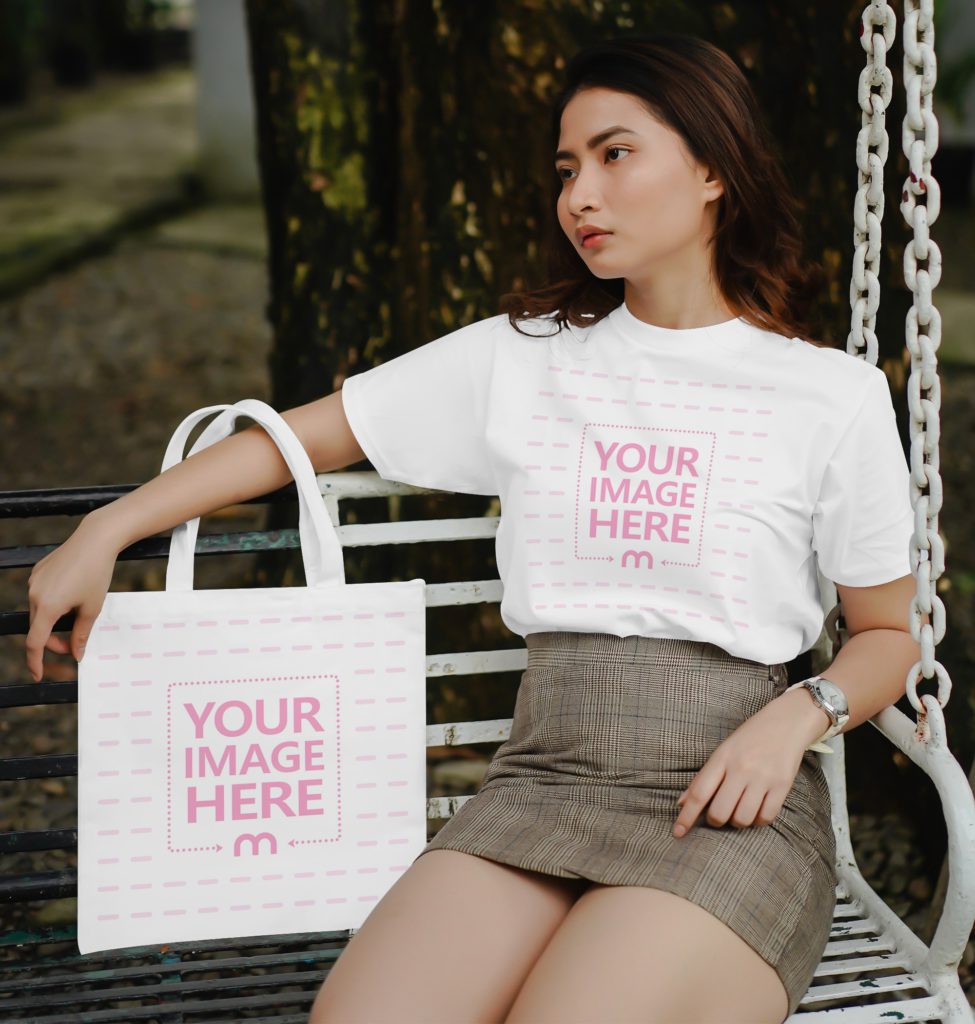 A woman is sitting on a park bench with a tote bag in her hand. He looks into the distance. She is wearing a t-shirt and a skirt.