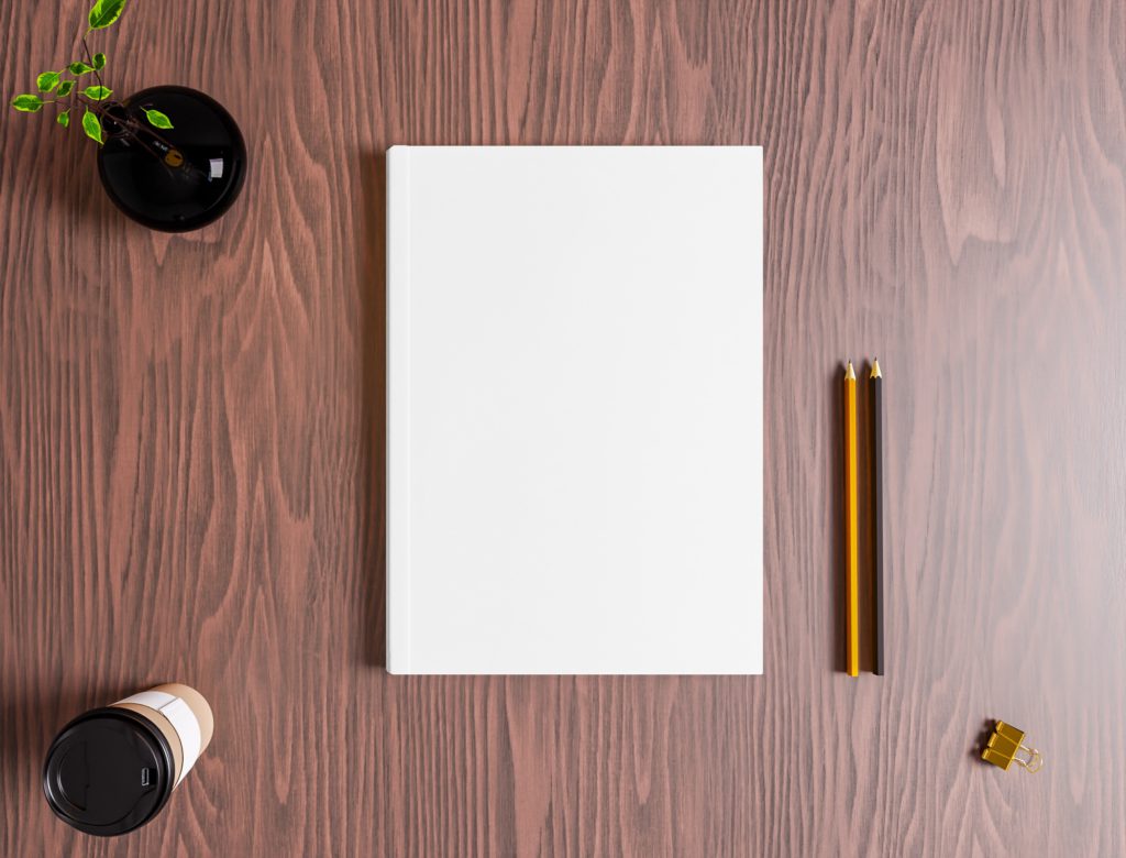 The notebook is on a brown table with pens, a plant and a cup of coffee.
